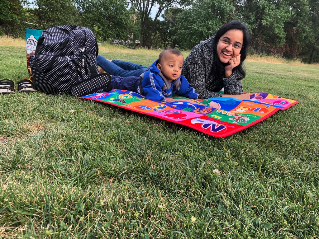 Mom and baby lying on her tummy on a playmat. The playmat is on the grass with a diaper bag by the side.